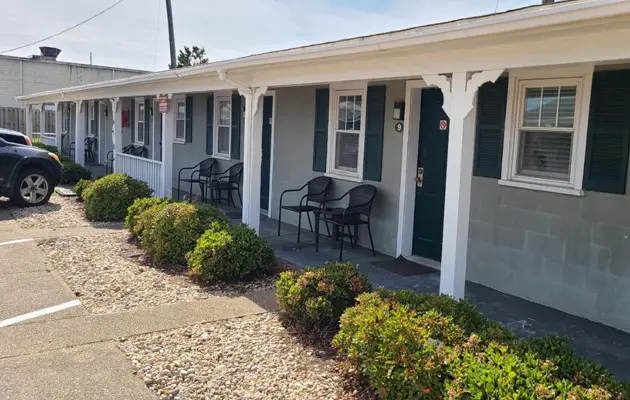 The covered porch along the front of the motel, with rocking chairs, potted shrubs and cars parked outside