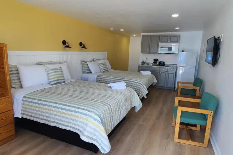 A guest room with two beds in cream and gold striped bedding, a kitchenette and two teal chairs beyond