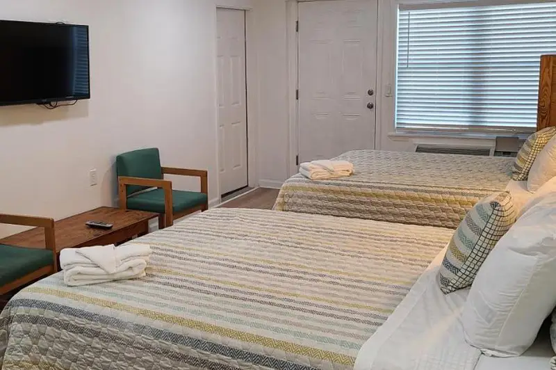 A guest room with two beds in cream and white bedding, a wall-mounted TV and green armchairs