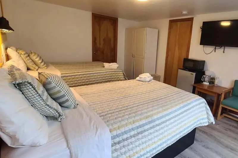 A guest room with two beds in cream and gold striped bedding, a wall-mounted TV and an armchair